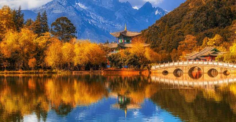 Pagoda tradicional y puente de piedra arqueado reflejados en un estanque cristalino con árboles de follaje otoñal amarillo y montañas nevadas al fondo.