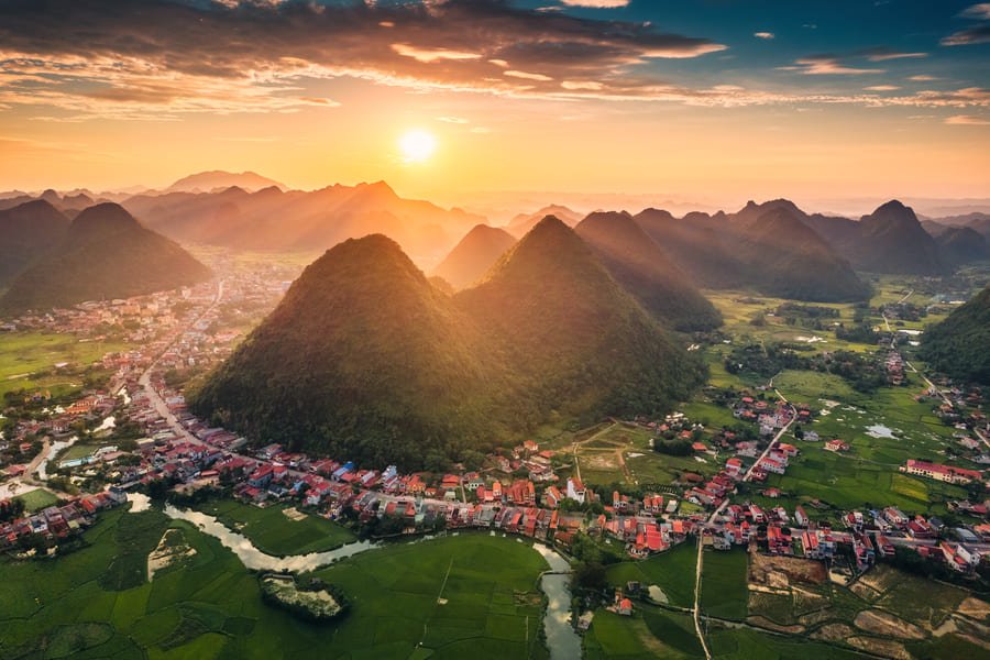Vista aérea del Valle de Bac Sơn en Vietnam durante un atardecer radiante. El sol poniente baña con luz dorada majestuosas montañas kársticas con forma de cono. A sus pies se extiende un río sinuoso, campos agrícolas verdes y un pintoresco pueblo rural con techos rojizos bajo un cielo cubierto de nubes iluminadas